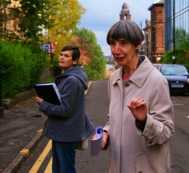 Colour photo of guides Joyce and Esther, standing on a leafy street with red sandstone building in the background. Esther is turned towards the camera, sharing information with a group of people who are out of sight.