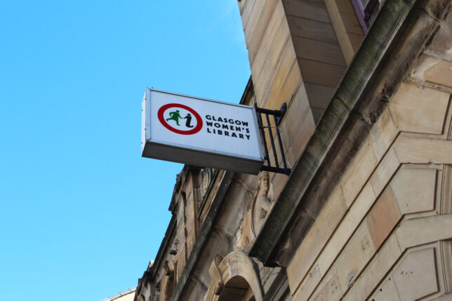 GWL Building Sign featuring the GWL logo of a red circle with a green figure of a walking woman and an 'i' for information, beside the text Glasgow Women's Library. The sign sticks out from the blonde sandstone walls of the building, and the sky above is blue.