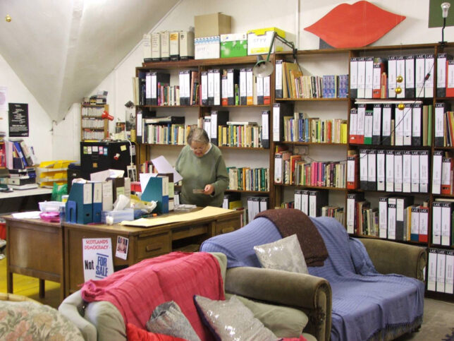 The Lesbian Archive set up in the Trongate space - a wall of shelves full of books and magazine holders, with a desk and two old sofas in front
