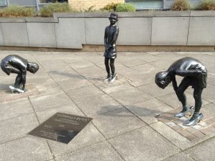 Photo of the "Gorbals Boys" on the corner of a street: three life-size bronze statues of three boys in silver high heel. Two of the boys are bent over as if to buckle up the shoes. The third is standing, holding his hands together at the groin. There is a plaque in the ground with the names of the boys: Joe, Nicky and Lee