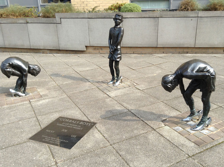 Photo of the "Gorbals Boys" on the corner of a street: three life-size bronze statues of three boys in silver high heels. Two of the boys are bent over as if to buckle up the shoes. The third is standing, holding his hands together at the groin. There is a plaque in the ground with the names of the boys: Joe, Nicky and Lee
