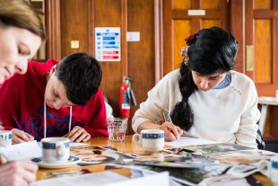 A small group sit round a table writing. Everyone has their heads down and looks to be focused. There are tea cups and what look to be photographic prompts on the table.