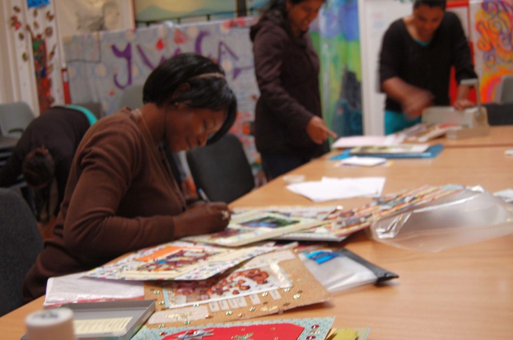 Three women sit around a table full of craft supplies making cards and badges. There are shelves full of books in the background.