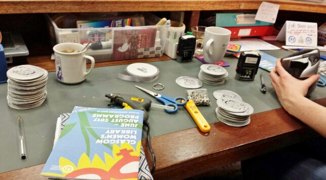 A table looking busy with scissors, mugs and other items scattered around. There are also small stacks of silver circles which seem to be the focus of the work.
