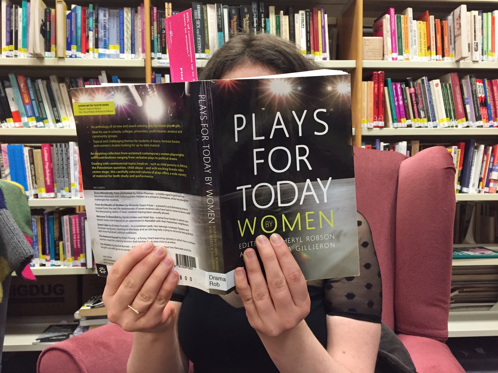 Photograph of person reading a book entitled 'Plays for Today: Women'. The person is sitting in a cosy chair in GWL with shelves of books visible in the background.