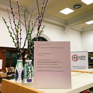 Photograph of new publication sitting on the top of GWL shelves beside decorations in the suffragette colours, purple, green and white. The publication is quite a large hard back book in a soft pink colour.