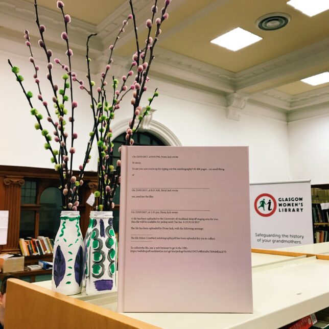 Photograph of new publication sitting on the top of GWL shelves beside decorations in the suffragette colours, purple, green and white. The publication is quite a large hard back book in a soft pink colour.