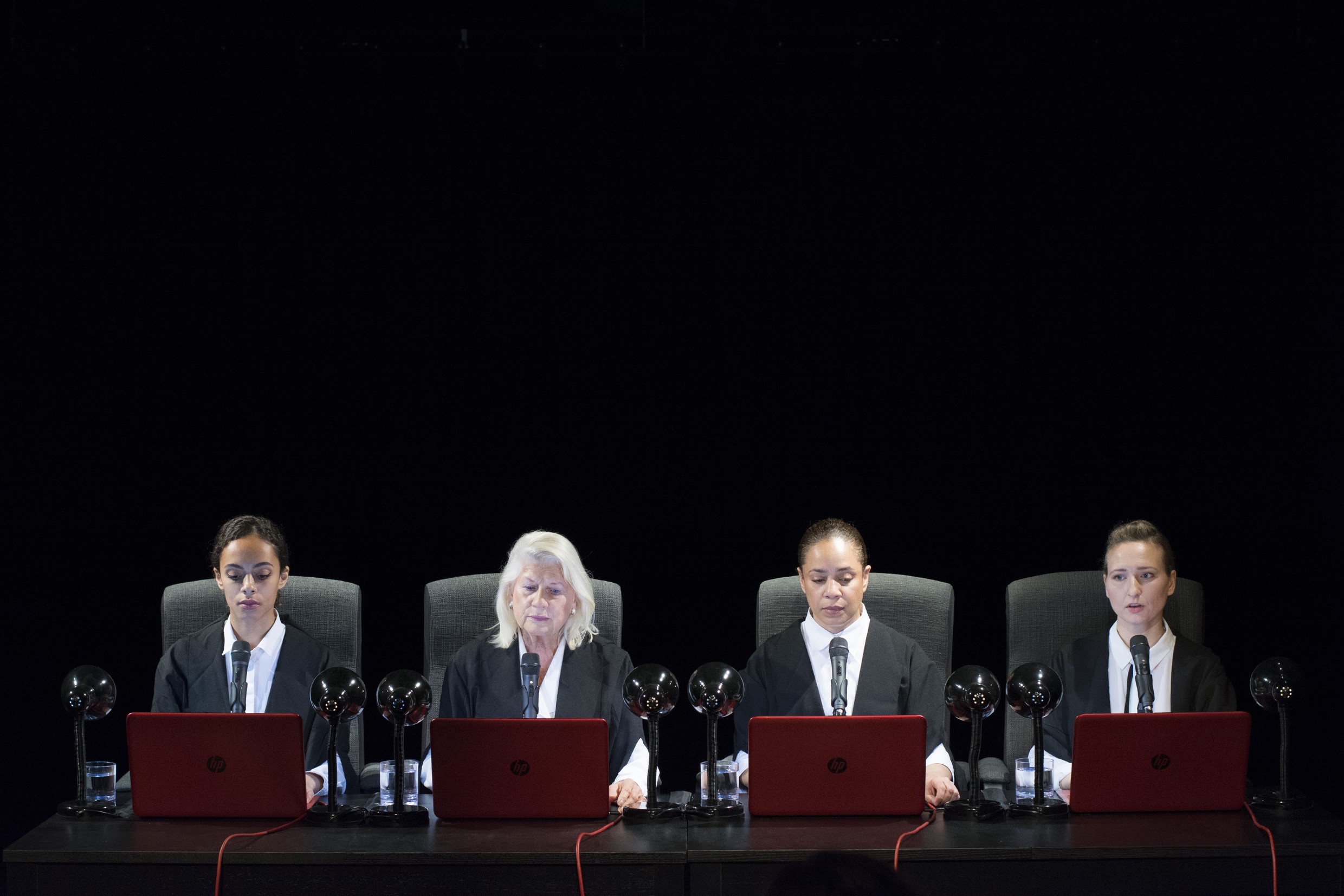 Photo of four women wearing suits sitting at a long desk. The background is black and the image is dark.