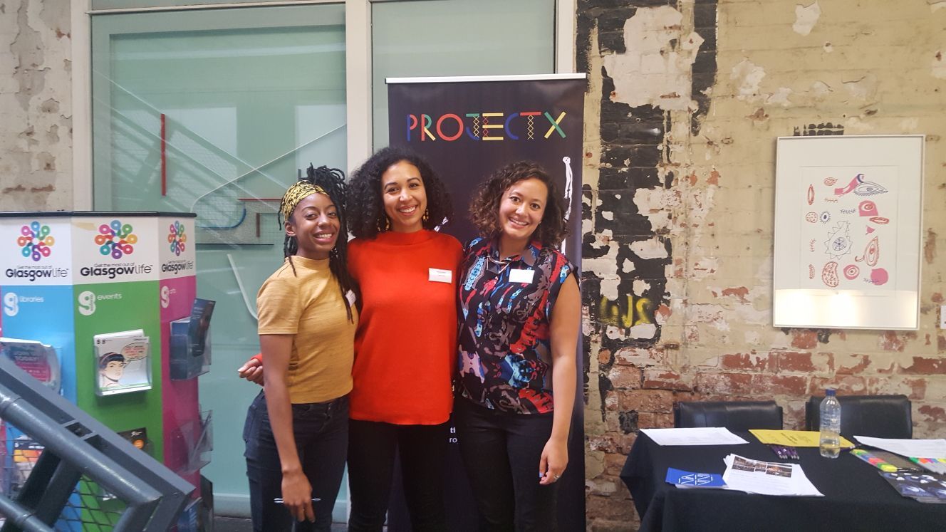 Photograph of three women of colour smiling and with their arms round one another. They are standing in front of a banner for 'Project X'.