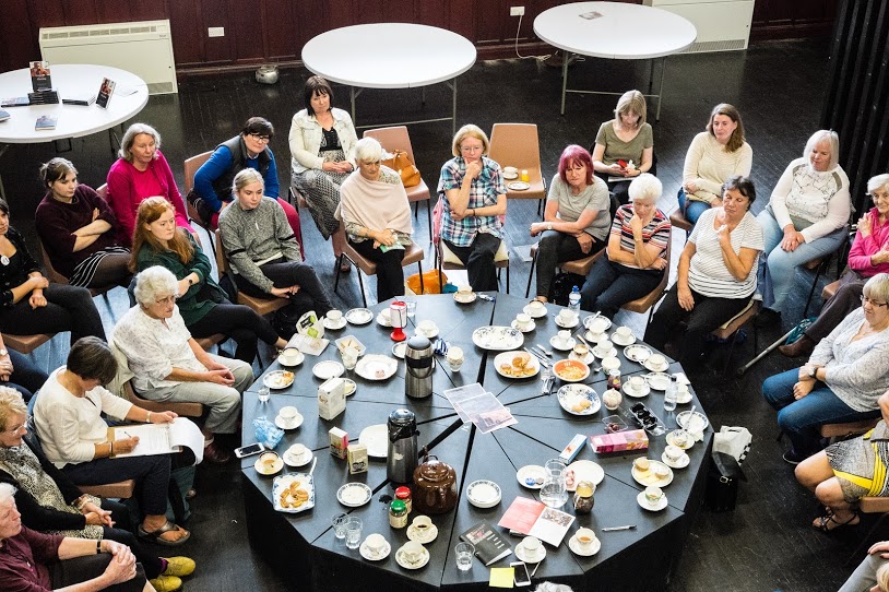 A large group of women sit round a big table covered with tea and food.