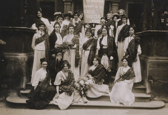 Black and white photograph of a group of suffragettes gathered on a set of stairs. Many of the suffragettes are wearing tartan sashes.