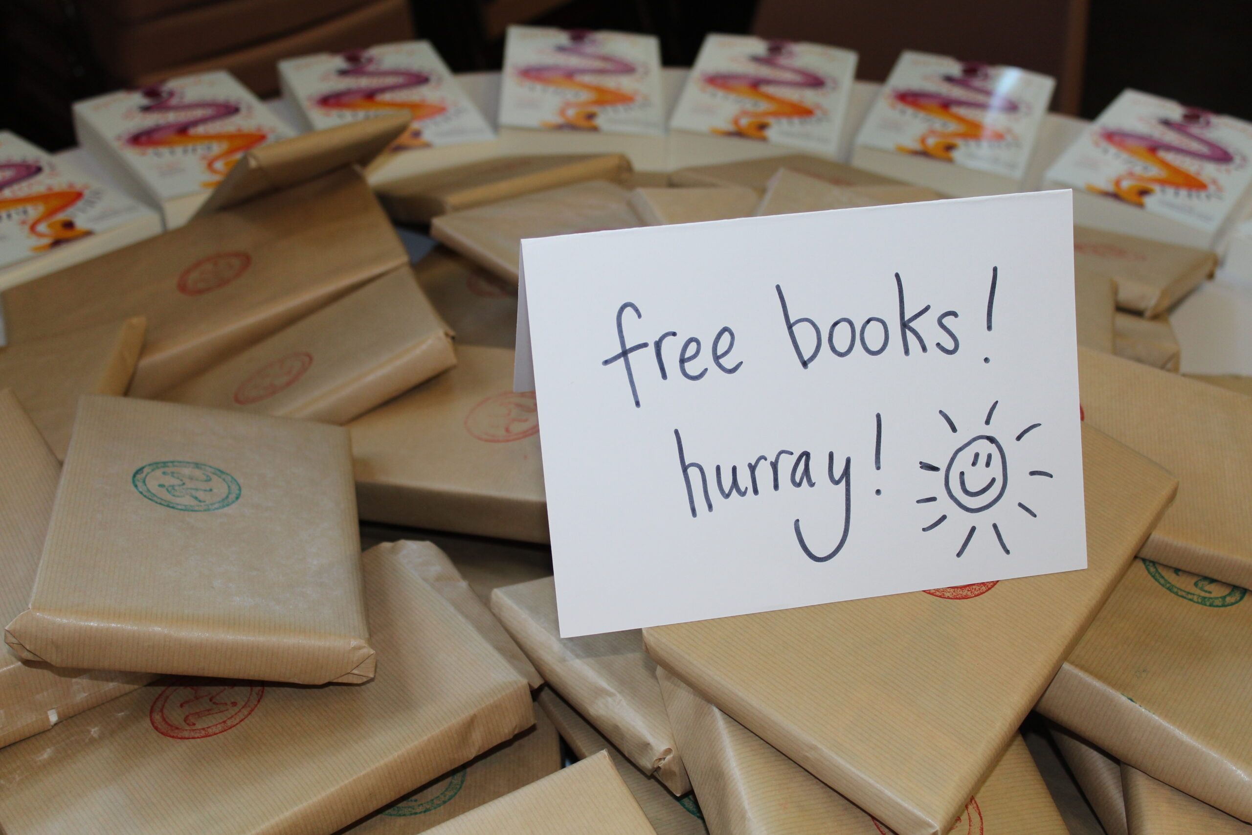 A big pile of books wrapped in brown paper with a handwritten label that says 'free books! hurray!' and has a line drawing of a smiling sun on it.