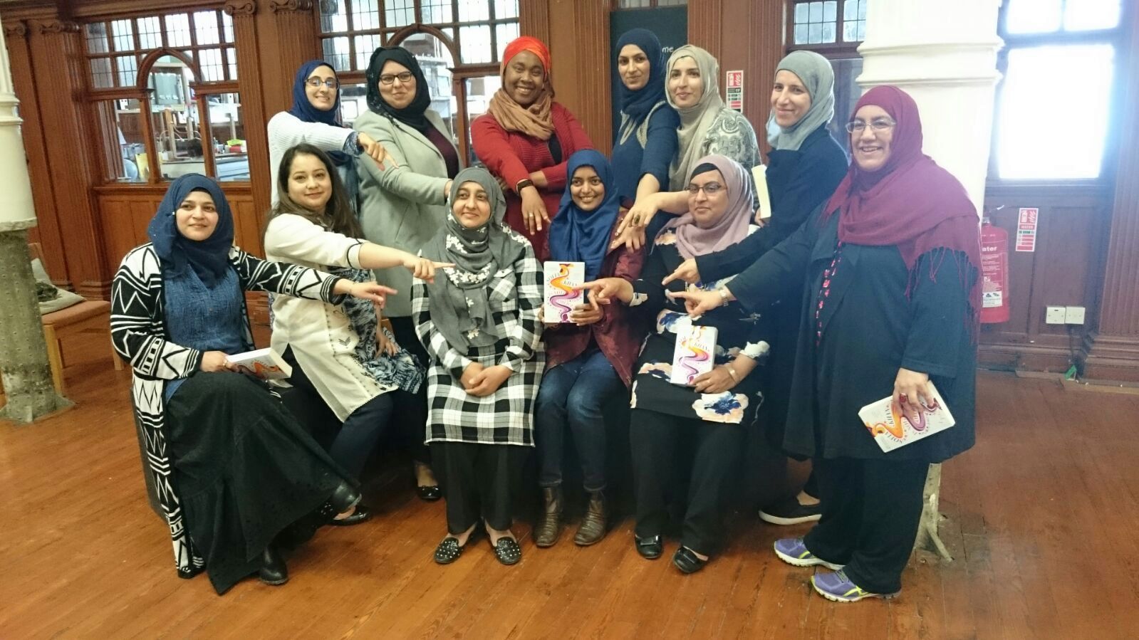 A group of women sit together and point at the book 'Is Not Obliged' by Sofia Khan which the woman in the middle is holding.