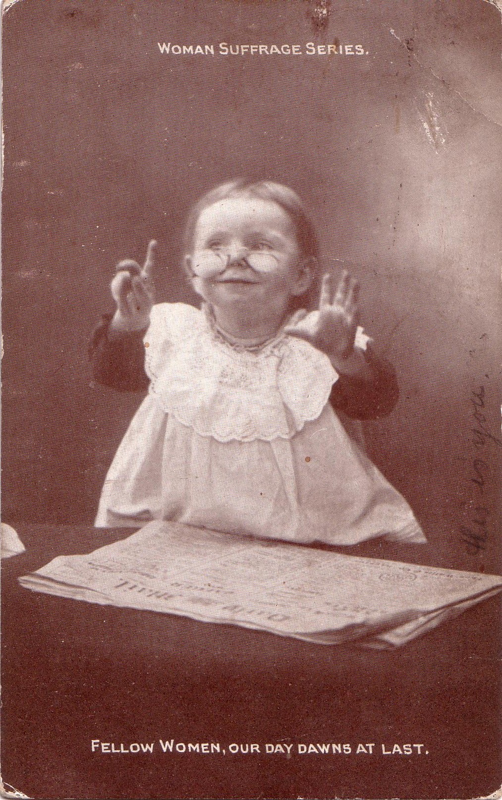 Black and white image of a young girl sitting at a desk in a school setting. Her eyes look inquisitive and she is raising her hand as if to answer a question.