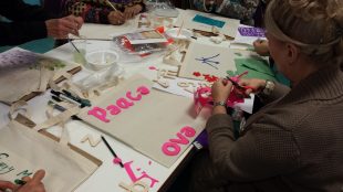 Women's hands making at a busy table, cutting out letters in pink and green to read 'Peace'.