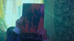 Image showing someone holding an old magazine in front of their face perhaps closely inspecting the cover or hiding. The room they are in has soft lighting with green tones from the curtains shining from behind them. 