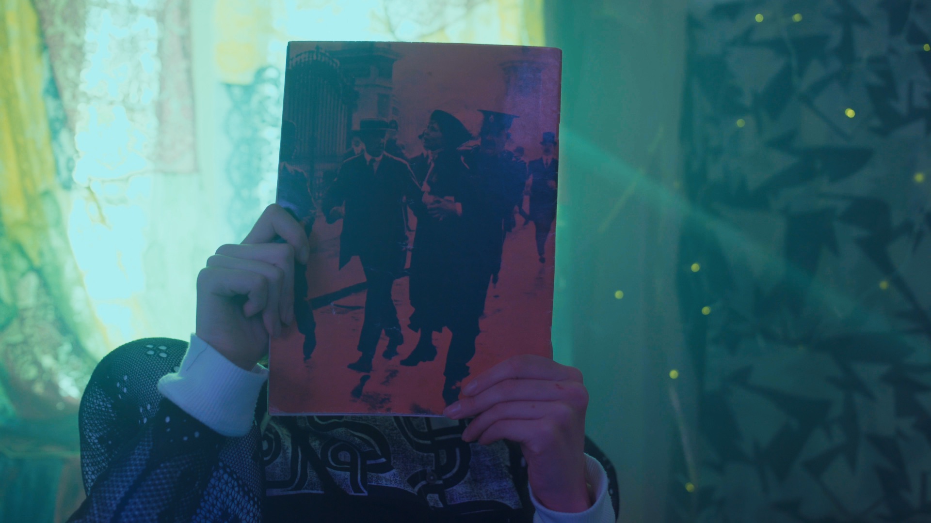 Image showing someone holding an old magazine in front of their face perhaps closely inspecting the cover or hiding. The room they are in has soft lighting with green tones from the curtains shining from behind them.