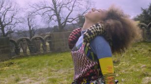 A woman leans her head back as if taking in the sunlight on her face or stretching back mid motion. There is an old stone wall in the background and she is in a field.