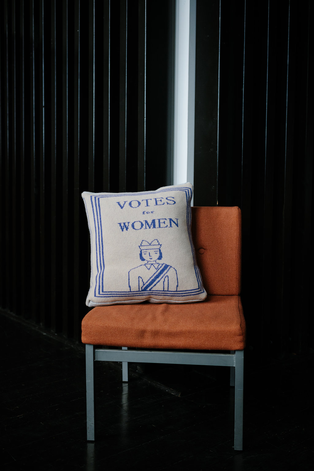 Donna Wilson's Book Cushion on a chair. The cushion has a knitted pattern with 'Votes for Women' above the image of a woman wearing a sash.