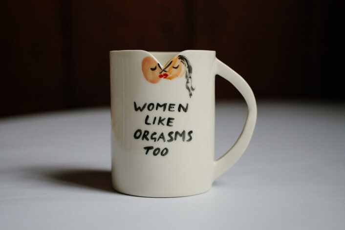 Women Like Orgasms Too Mug. At the top of the mug there are two figures kissing and here there is a small triangle cut out of the rim of the mug.