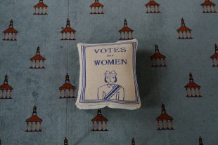 Image of a cushion with a knitted pattern with 'Votes for Women' above the image of a woman wearing a sash.