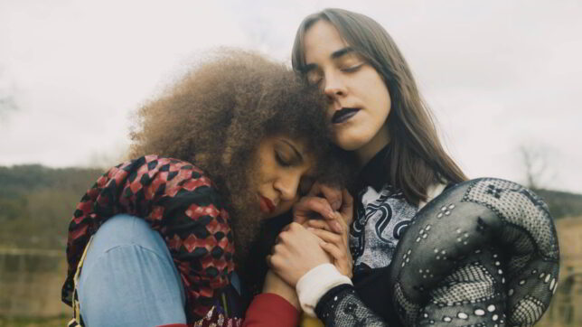 Image of two women holding hands and leaning their heads against one another. Their eyes are closed and they are wearing costumes that look like a modern spin on Tudor fashions with puffy sleeves and lace.