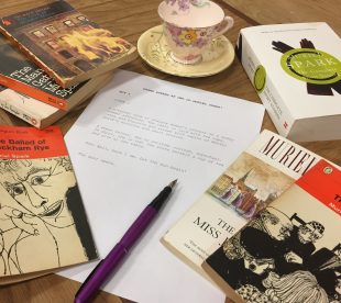Photograph of a table of Muriel Spark novels surrounding a piece of paper with the beginnings of a script written about a women coming to the library for Drama Queens.
