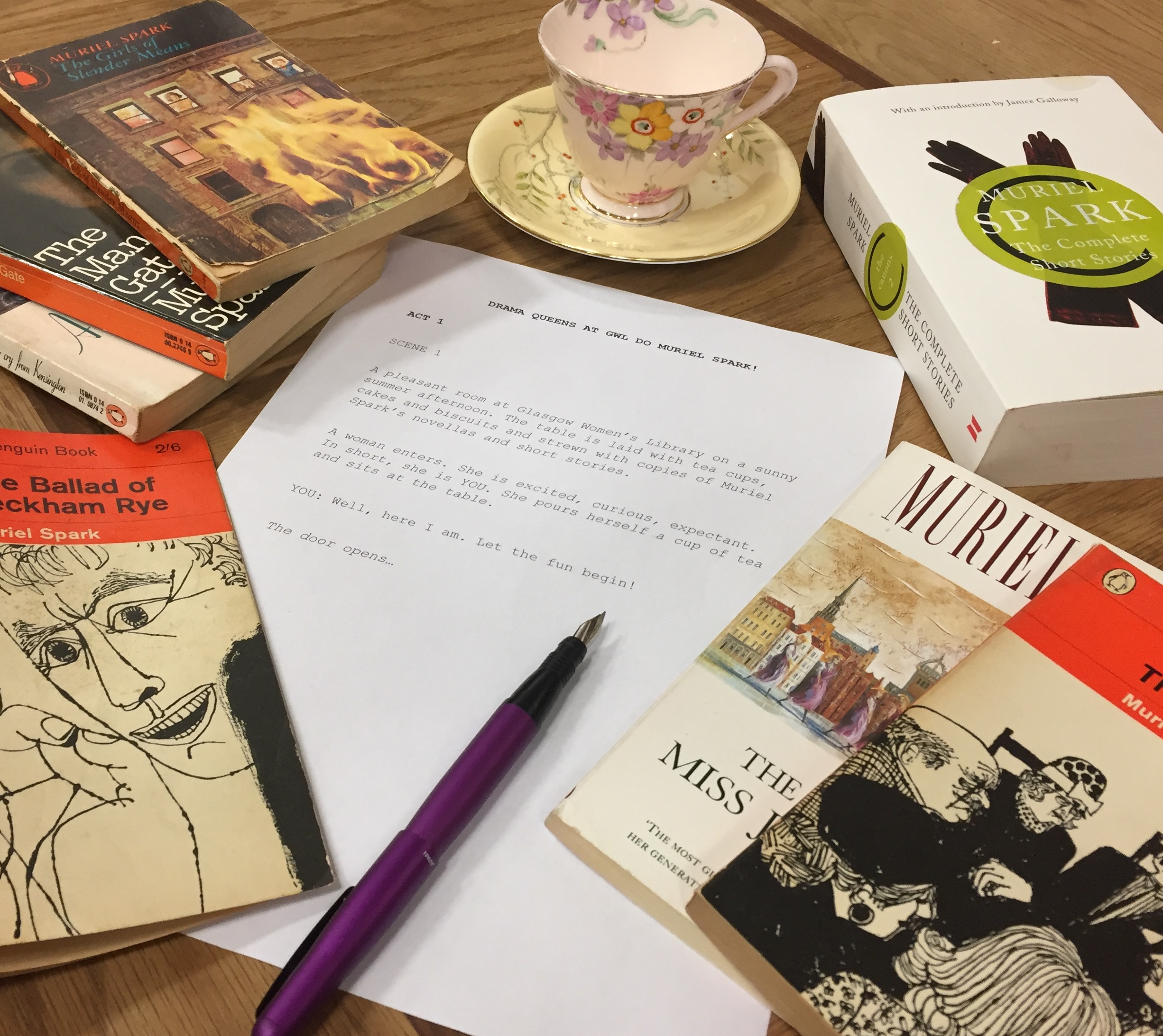 Photograph of a table of Muriel Spark novels surrounding a piece of paper with the beginnings of a script written about a women coming to the library for Drama Queens.