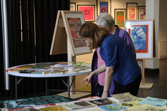 Two women stand looking at materials that are laid out on tables,