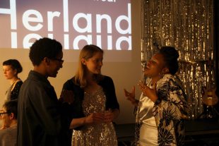 Three women stand in front of a projection that says 'Herland'. They are talking animatedly and look happy