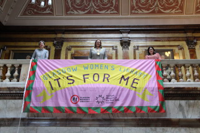 Three woman standing holding a flag that drapes over a marble balcony. The flag is pink with a yellow banner and the words 'Glasgow Women's Library' and 'It's for me' are written on the flag.