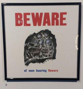 A framed print with a central image of a hand holding some blue flowers. The text above the image reads BEWARE in red capital letters; the text below reads 'of men bearing flowers', in blue and red font. The quote is from Muriel Spark's autobiography, Curriculum Vitae.