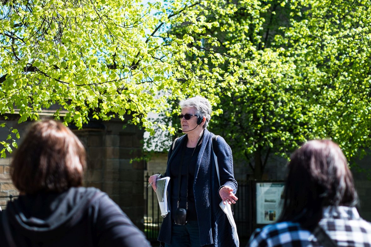 Tour guide Anabel is speaking to a group. It’s a sunny summer day and there are bright green trees behind her.