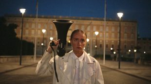 Film still showing a woman in a white jacket standing outside at twilight holding a megaphone in her right hand. In the background is a building and she is standing in the middle of an empty street or drive way.