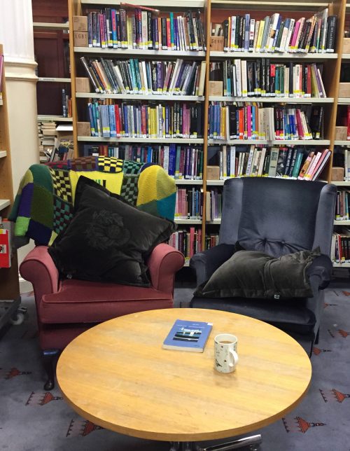 An image of our comfy chairs in the Library space, The shelves in the background are full of books. The table has a book and a mug on it. The left hand chair has a pillow on it. The right hand chair has a pillow on it that has fallen down.