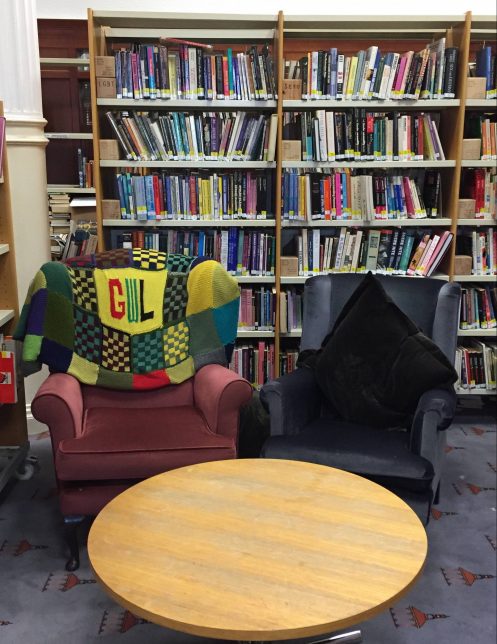 An image of comfy chairs in our library. The table in front of the chairs has nothing on its surface. The left hand hair has no pillow on it. The right hand chair has a pillow that has been propped up.