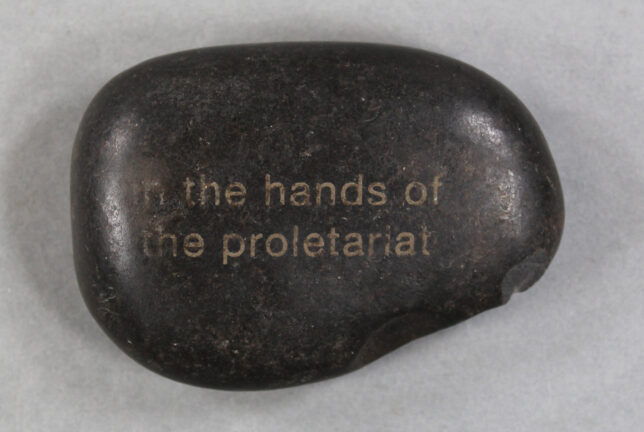 A cobblestone from Fiona Jack's exhibition Our Red Aunt. The stone is smooth and dark, just the right size to fit in your hand, and has the phrase 'in the hands of the proletariat' engraved on the surface.