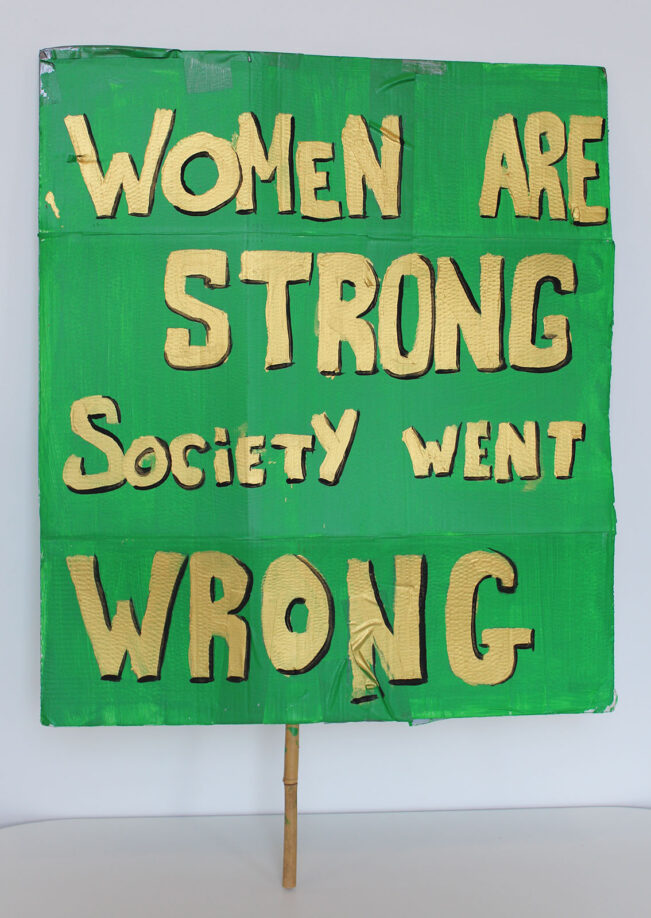 Green placard reading 'Women are strong, Society went Wrong' in strong gold lettering