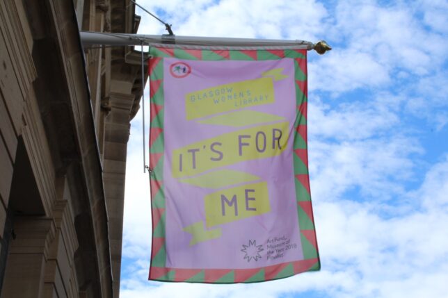 Flag designed by Visual Communication Resident (2018) Jyni Ong for our Art Fund Museum of the Year public engagement - on a pink background with a bold red and green border, a twisting yellow ribbon reads: Glasgow Women's Library: It's For Me