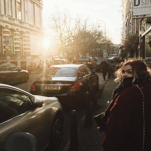 Photograph of Mhairi walking down the busy streets of Amsterdam, Netherlands. Mhairi is looking back into the camera. The sun is shining brightly into the camera reflecting the rays. Two cars are beside her parked. 