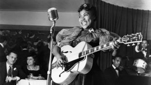 Sister Rosetta Tharpe with large guitar and classical condenser microphone. She is smiling. And she is in the middle of a performance at a club. 