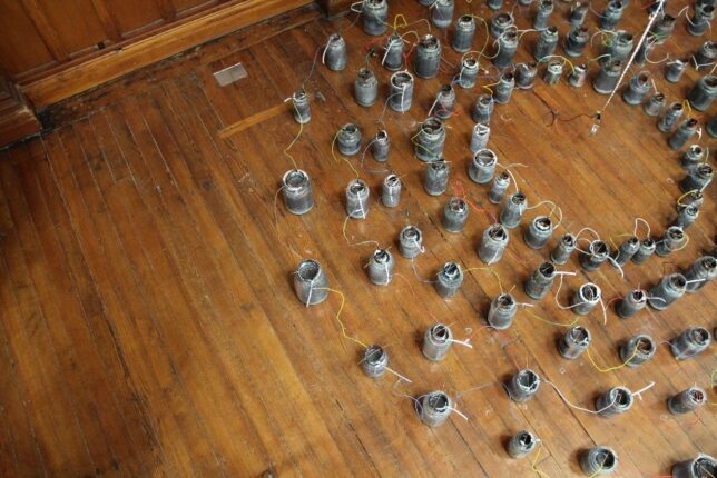 Concentric circles of mismatched glass jars on a wooden floor. The jars have salt crystals near the rim and are connected with wires, and a light strip hangs at their centre.