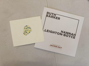An exhibition catalogue with the names Ruth Barker and Hannah Leighton-Boyce sits on a white table. On top of the catalogue is a postcard. The postcard is an off-white and in the centre are lots of small paper sqaures grouped together. The squares are yellow and green and together look like the shape of a pear.