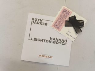  An exhibition catalogue with the names Ruth Barker and Hannah Leighton-Boyce sits on a white table. On top of the catalogue is a postcard. On the right hand side of the postcard is a big bow made of black ribbon, below it are the handwritten words 'Lady, enjoy your life.' On the left side of the postcard is a pink piece of paper on which a stamp has been used, the stamp is a long list of words including 'joy' 'dream' 'perfect' and 'love'.