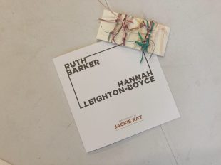 An exhibition catalogue with the names Ruth Barker and Hannah Leighton-Boyce sits on a white table. On top of the catalogue is a postcard. The postcard is an off-white colour and rectangular in shape. Tied around the postcard are lots of thin overlapping ribbons in different colours.