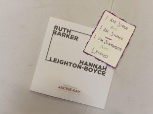 An exhibition catalogue with the names Ruth Barker and Hannah Leighton-Boyce sits on a white table. On top of the catalogue is a postcard. The postcard is off-white with a purple edge. In the centre are the handwritten words 'I am siren and I am silence. I am suffragette and legend'. The words are purple and green.