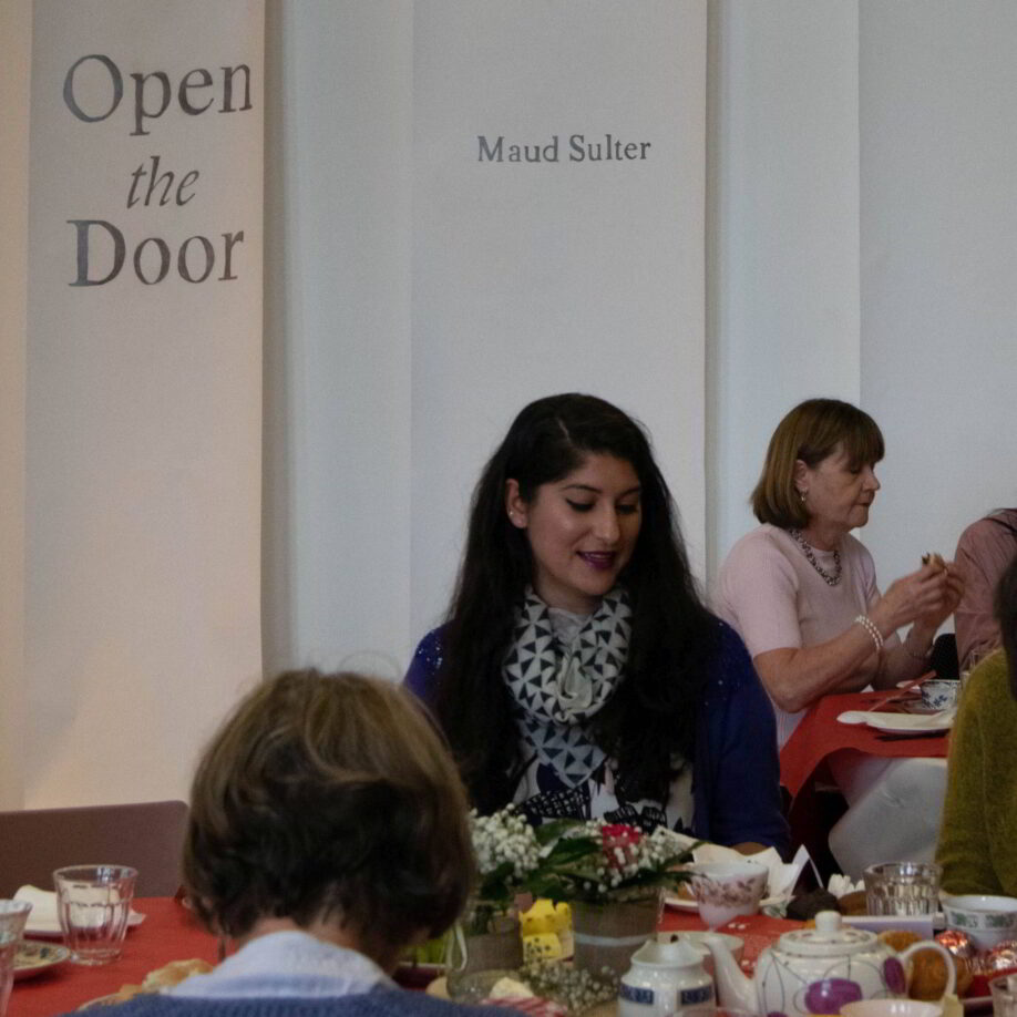 Women sitting around a table with teapots and cakes on it. Behind them is a banner that has 'open the door' cut out of it.
