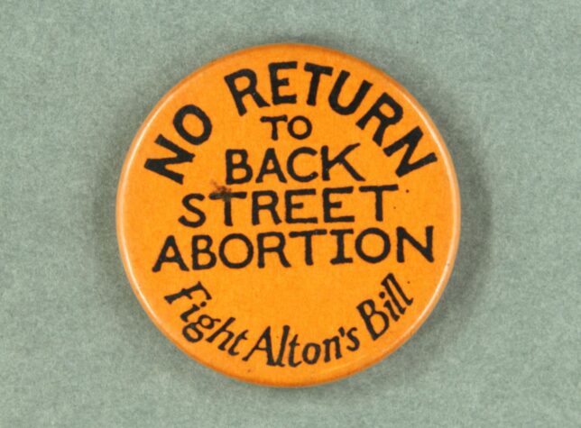 An orange badge that has black text that reads, "No Return to Back Street Abortions, Fight Alton's Bill".