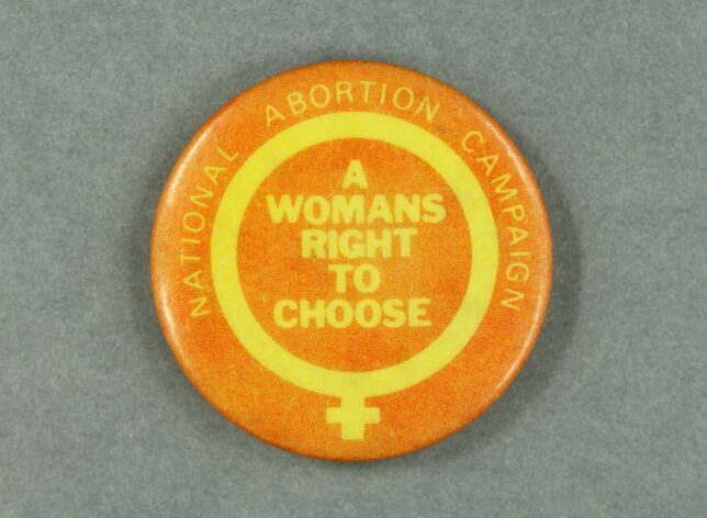 An orange badge with a yellow symbol for 'woman' on it. In the centre of the symbol is the yellow text, "A Womans Right to Choose".