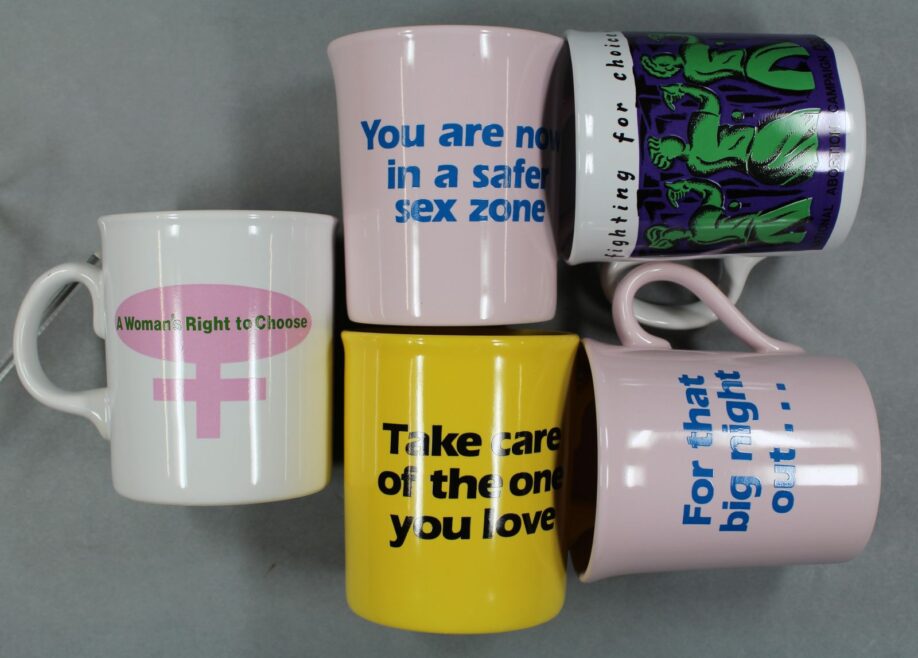 Five mugs arranged in a cluster. One reads, "You are in a safer sex zone" and another is a pro-choice mug from the National Abortion Campaign.